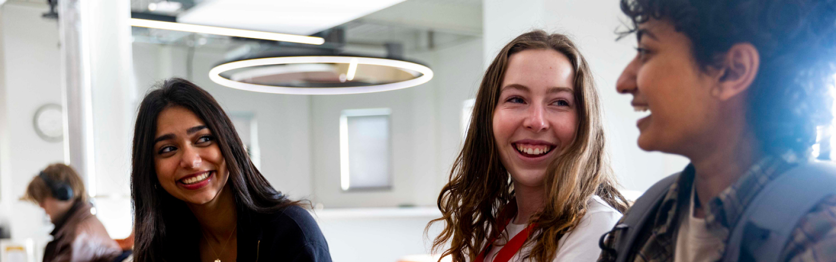 Three people are sitting together indoors, smiling and talking. They appear to be in a modern office or study space. A circular ceiling light is visible above them.