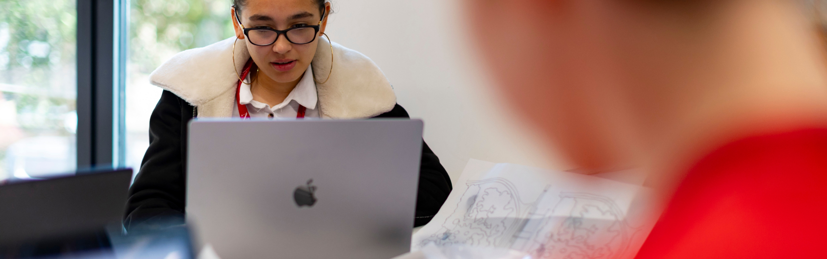 A person wearing glasses and a jacket works on a laptop at a desk. Papers and a blurred person in a red shirt are visible in the foreground. Natural light comes through a window in the background.