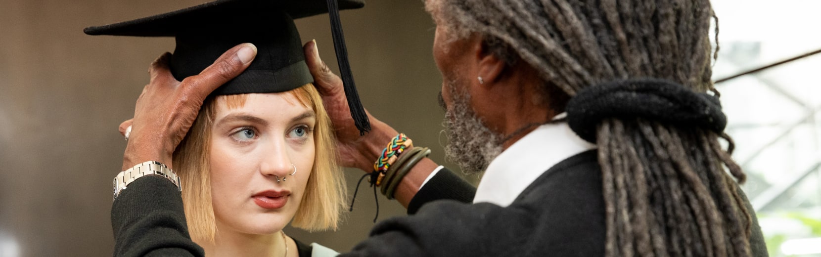 An older person adjusts the graduation cap of a young adult who is wearing a nose ring and has short blonde hair. The scene suggests a graduation ceremony or special moment of achievement.