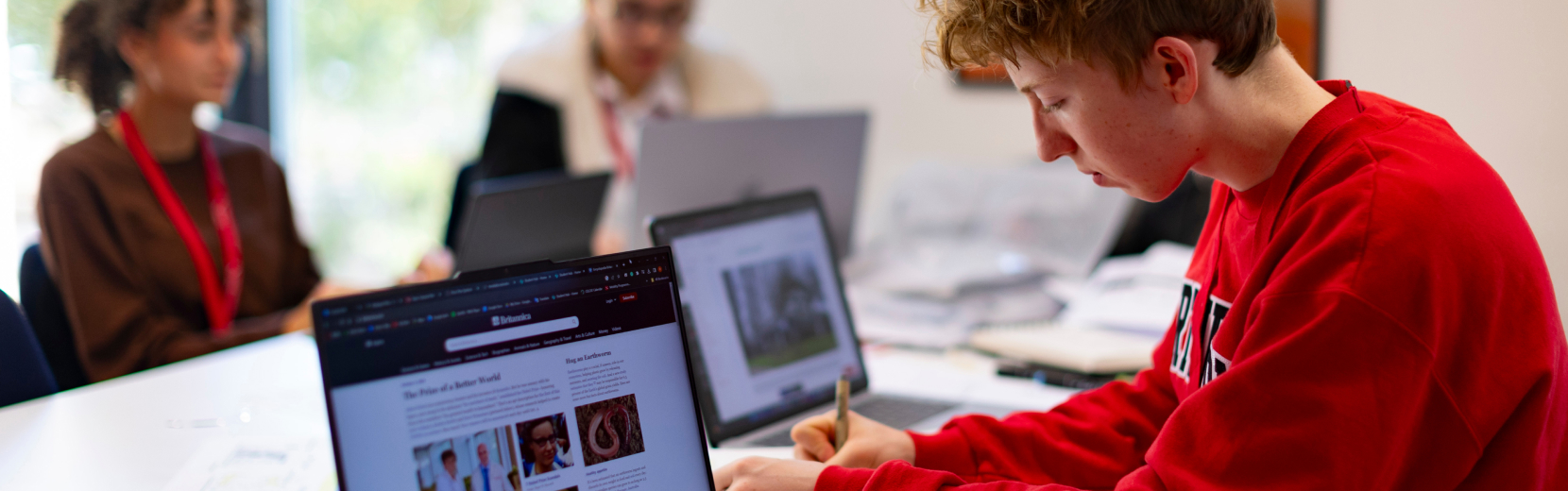 A group of students are sitting at a table with laptops and notebooks. One student in a red sweatshirt is writing on a notepad, while others are focused on their laptops. The scene suggests a collaborative study session.