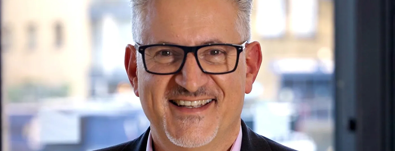 A man with gray hair and glasses is smiling. He is wearing a dark blazer and a light-colored shirt. The background is softly blurred, showing large windows with an urban view outside.