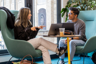 Two people sit in teal chairs at a small table with laptops and a drink. The woman on the left gestures while engaging in conversation. They are in a modern setting with large windows and plants in the background.