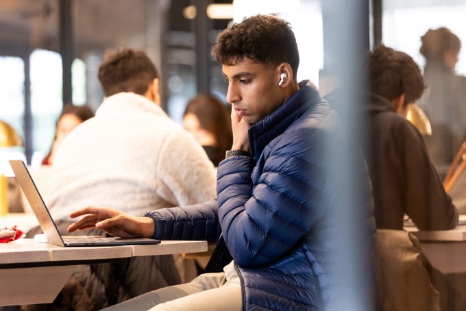 A person wearing a blue jacket and earbuds is working on a laptop in a busy indoor setting. Several other people are seated around, engaged in various activities. The scene suggests a shared workspace or café environment.