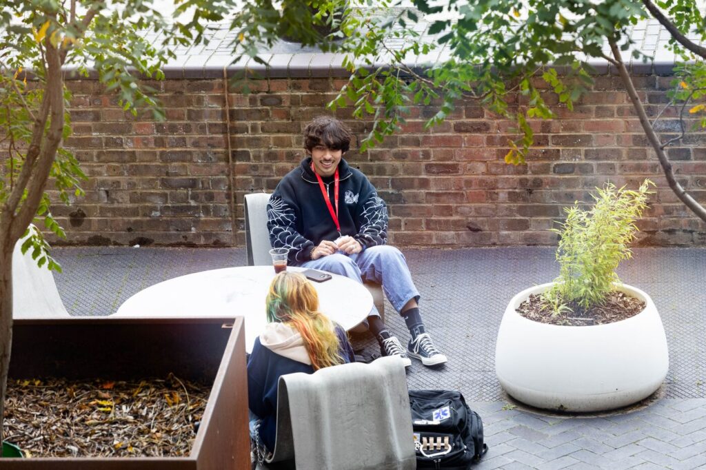 Two people sitting at a round outdoor table, engaged in conversation. One person, wearing glasses and a black jacket, appears to be smiling. Green plants are in large pots nearby, and there's a brick wall in the background.