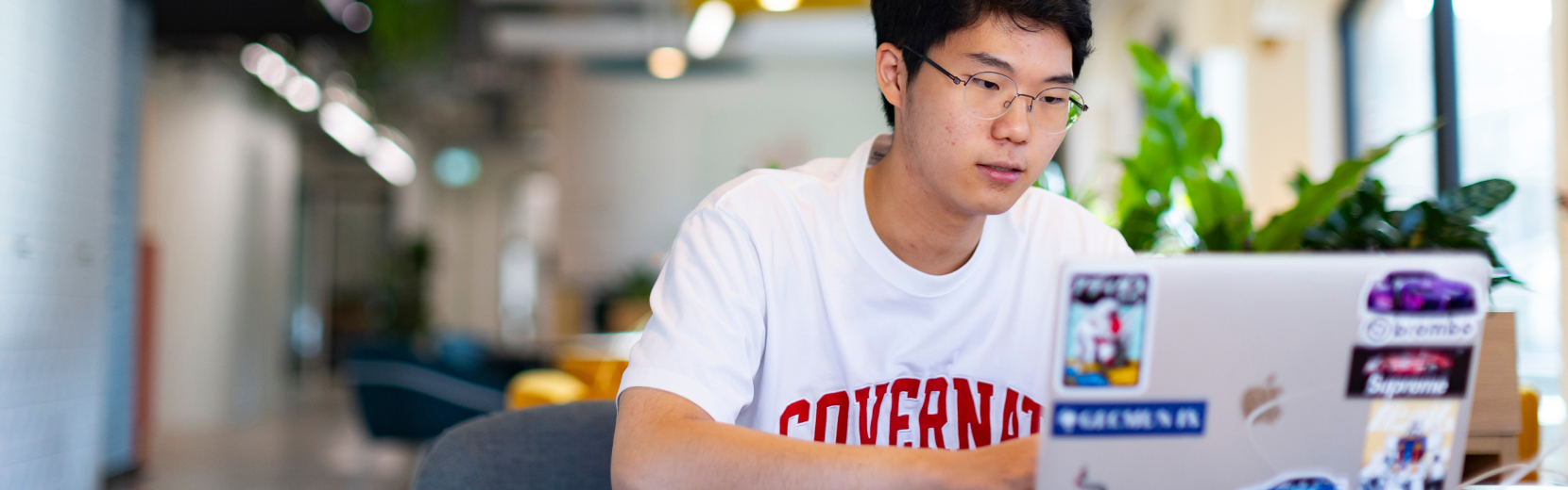 A person wearing a white t-shirt and glasses is focused on working at a laptop decorated with various stickers. The background features a modern, brightly lit office space with plants and seating areas.