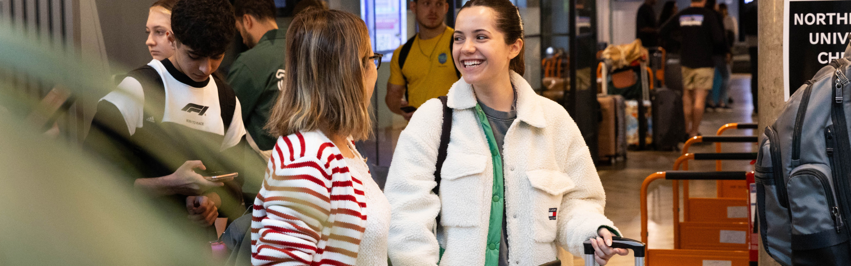 A woman in a white jacket smiles while talking to another woman wearing glasses and a striped top. They are in a busy indoor area with people in the background, some holding luggage.