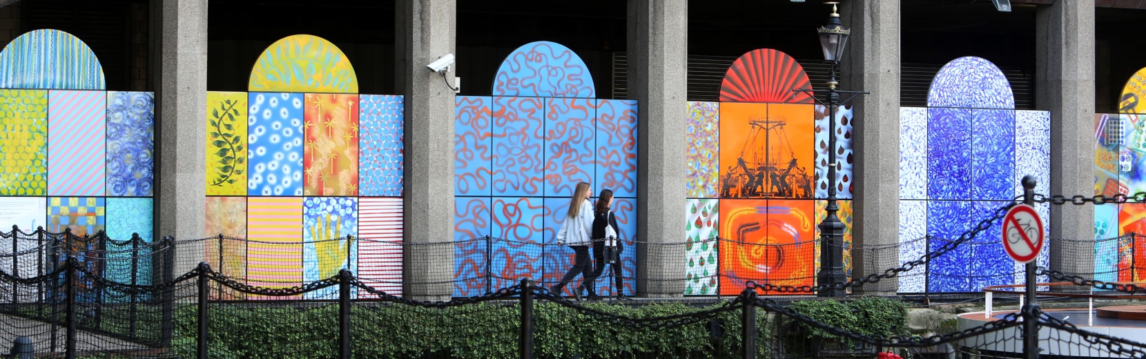 A person walks in front of a series of colorful, abstract murals displayed on arch-shaped panels. The murals feature various patterns and designs, including wavy lines and geometric shapes, set against a concrete building with columns.