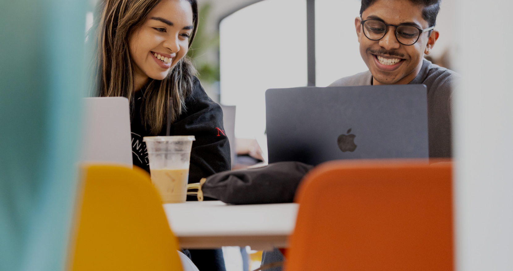 Two people sitting at a table with laptops, smiling and working together. There's an iced coffee on the table, and the chairs are orange and yellow, creating a bright and cheerful atmosphere.