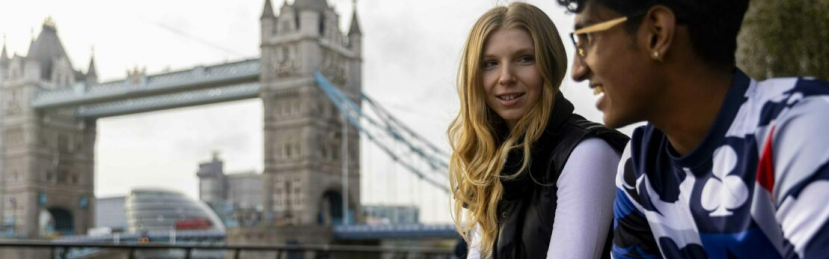 Two people sit and smile near Tower Bridge in London. One wears a white shirt and dark vest, the other wears glasses and a patterned shirt. The bridge and city buildings are visible in the background.