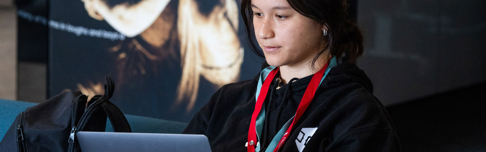 A person wearing a black hoodie and a red lanyard is focused on typing on a laptop. A black bag is placed beside them.