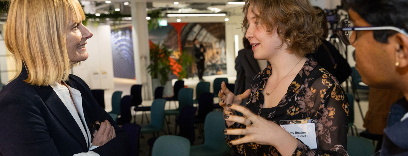 Three people are engaged in conversation in an office setting; one woman gestures while speaking. The background features chairs and abstract wall designs, creating a modern, professional atmosphere.