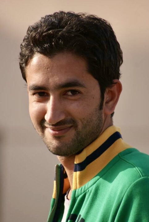 Dr. Anees Baqir, a smiling man with short dark hair, dons a green jacket accented by yellow and navy stripes on the collar, standing confidently against a neutral background.