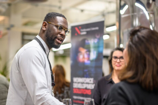 A man speaking to a woman at a professional event. They are in an office setting with a blurred promotional banner in the background. Both are dressed in business attire.