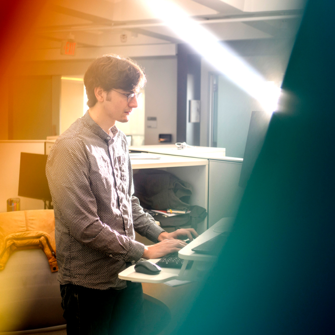 A person with glasses works at a standing desk in a modern office. Warm light filters through the room, creating a vibrant atmosphere. The workspace is organized and features computers and office equipment.