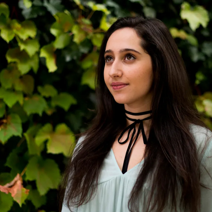 A woman with long dark hair stands in front of leafy greenery, looking upward. She wears a light blouse with a black ribbon tied as a choker.