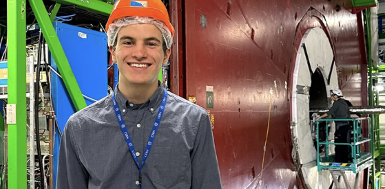 A person wearing a hard hat and lanyard smiles in front of a large industrial machine with red and white components. Another worker in safety gear is visible on scaffolding in the background.