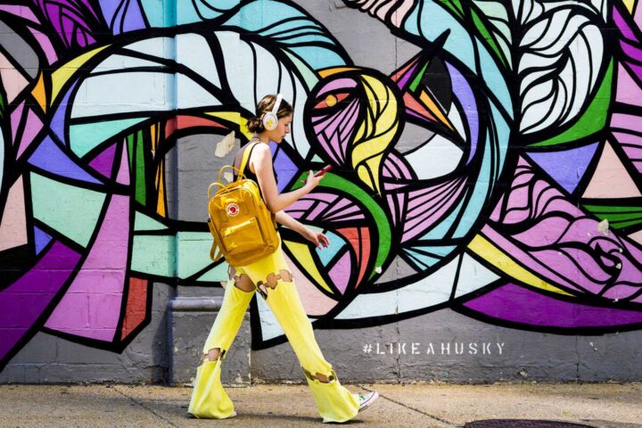 A person wearing yellow pants and a yellow backpack walks past a vibrant, abstract mural. They are listening to music with headphones. The mural features swirling patterns in various colors. Text on the sidewalk reads "#LIKEAHUSKY.