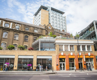 Street view of a modern and historic brick building with arched windows. People are walking and standing outside. A tall, contemporary building is visible in the background under a cloudy sky.