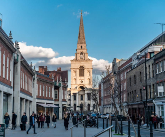 A city street scene with people walking on a pedestrian path. In the background, a large church with a tall steeple is visible under a blue sky with clouds. Buildings line both sides of the street.