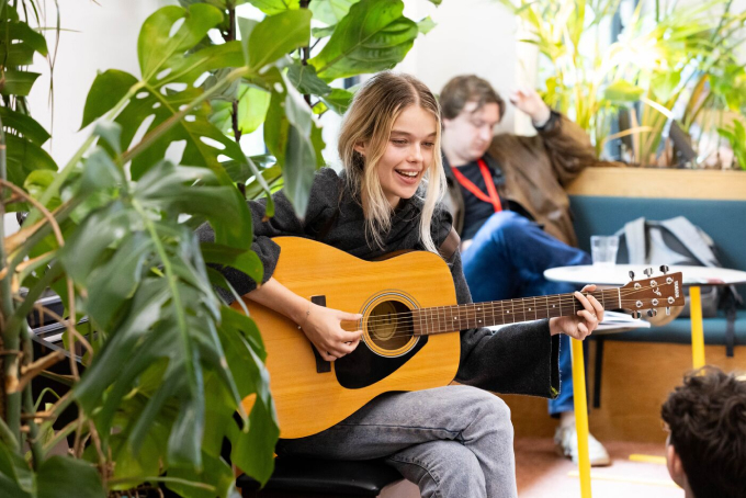 A woman sits among green plants, playing an acoustic guitar and smiling. She wears a black top and blue jeans. In the background, a man sits on a couch using a phone.