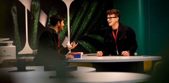 Two people are sitting at a desk having a lively conversation. Both are wearing red lanyards. The background features large images of green plant leaves.