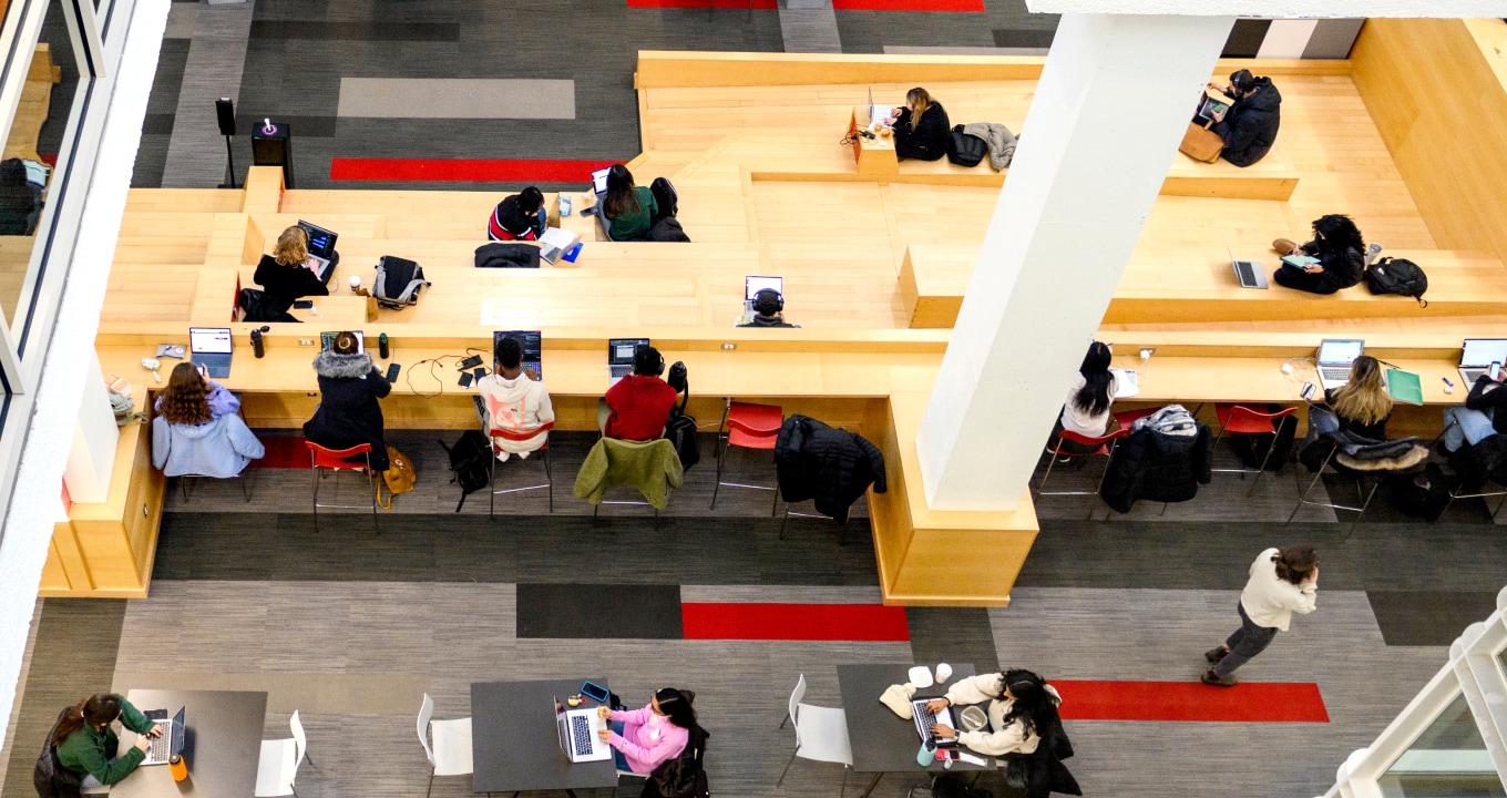 Bird's-eye view of a modern library or study space with people seated at wooden desks and tables, using laptops and studying. The flooring has a geometric pattern with gray and red accents.