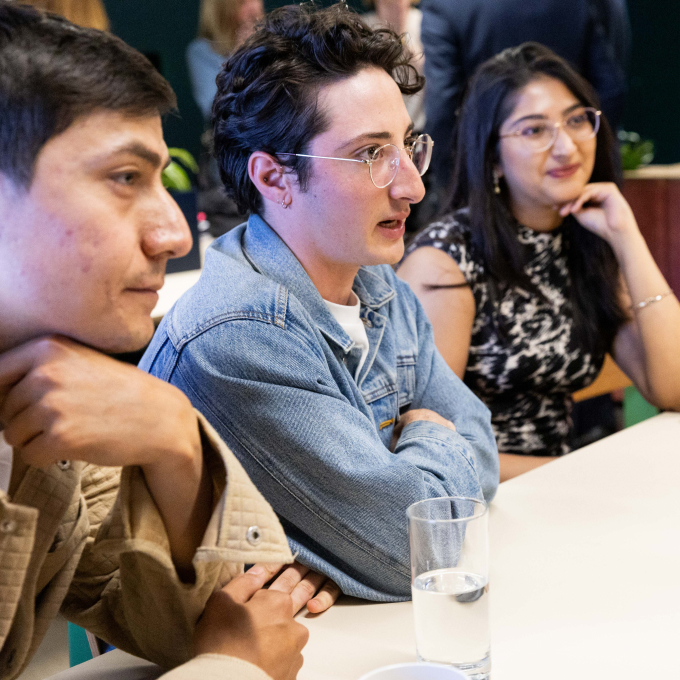 Three people sitting at a table engaged in conversation. One person wears a denim jacket, another a patterned dress, and the third a beige jacket. A glass of water is on the table. The background is slightly blurred.