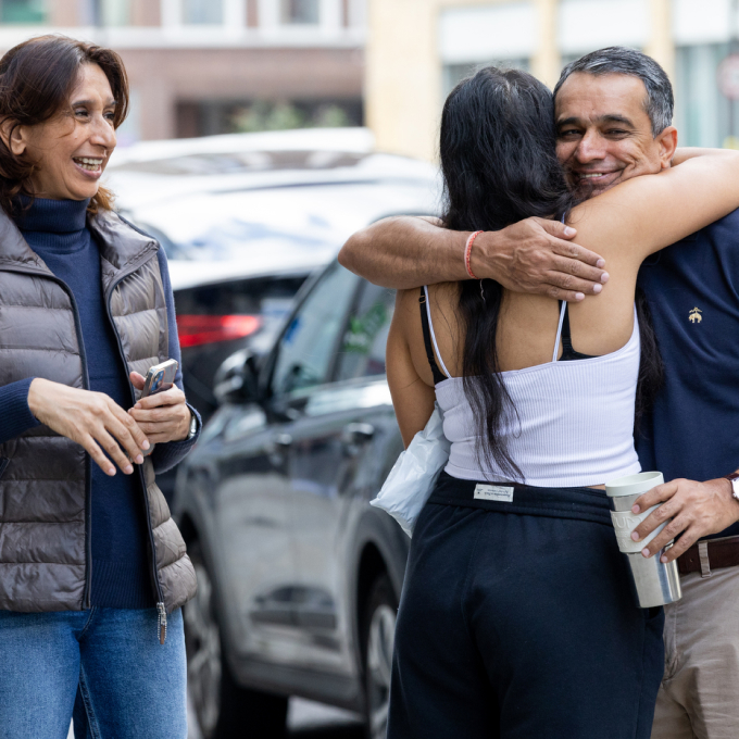 A man and woman hug warmly on a city sidewalk, both smiling. Another woman stands nearby, smiling and holding a phone. A parked car is in the background. It appears to be a happy, casual outdoor moment.