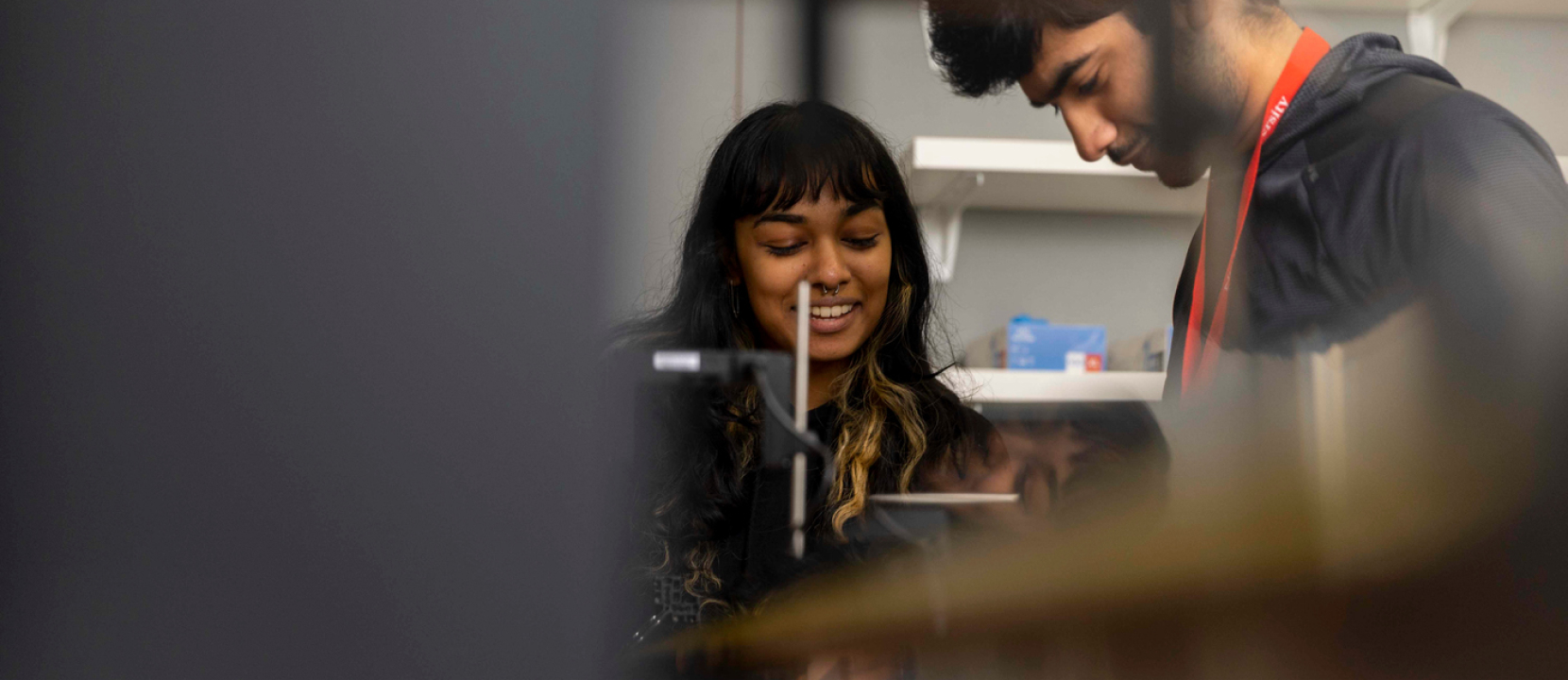 Two people are standing closely together, looking at a device. The person on the left is smiling, while the one on the right appears focused. Shelves with various items are visible in the background.