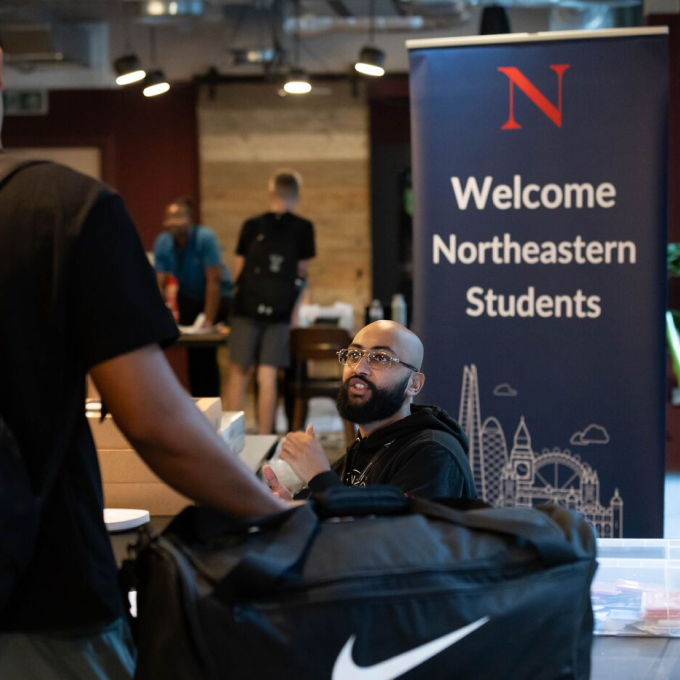 A bearded man sits at a table with a large duffle bag in the foreground. A banner nearby reads "Welcome Northeastern Students" with a stylized cityscape. Other people are blurred in the background.