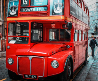 A classic red double-decker bus parked on a street, displaying the route number 15 to Tower Hill. The bus has an iconic design with vintage elements. A person walks nearby on the rainy pavement.