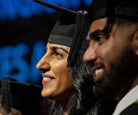 Two graduates in profile wearing black caps and gowns smile during a ceremony. The background is blurred, highlighting their expressions of joy and achievement.