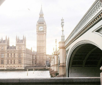 View of the Houses of Parliament and Big Ben in London, with the River Thames and Westminster Bridge in the foreground. The clock tower is prominently visible against a cloudy sky.