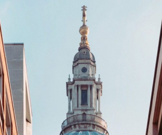 A tall, ornate dome with gold accents and a cross on top, framed by two modern buildings on either side against a clear blue sky.