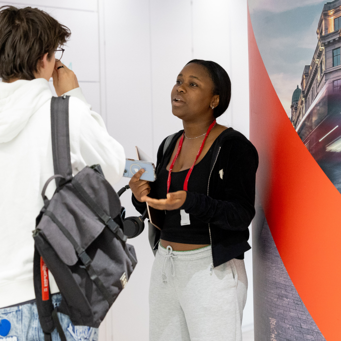 A woman in casual clothing, holding a phone, talks to a man with glasses and a gray backpack. They are standing indoors near a red and gray wall panel. The man adjusts his glasses while listening.