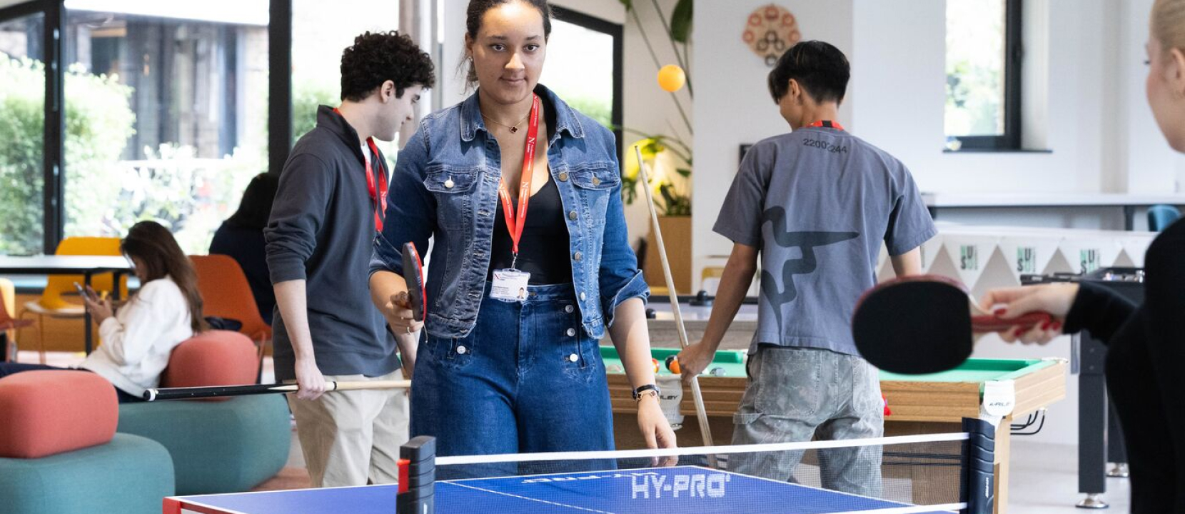 A group of people are playing ping pong in a recreational space with colorful furniture. The focus is on a woman in a denim outfit holding a paddle, while others are engaged in activities around her.