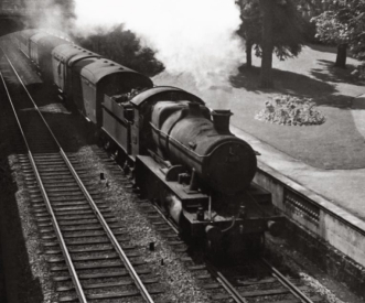 A vintage steam locomotive pulling a passenger train moves along railroad tracks. The train emits white steam into the air. Surrounding the tracks is a grassy area with trees and bushes, suggesting a countryside setting.