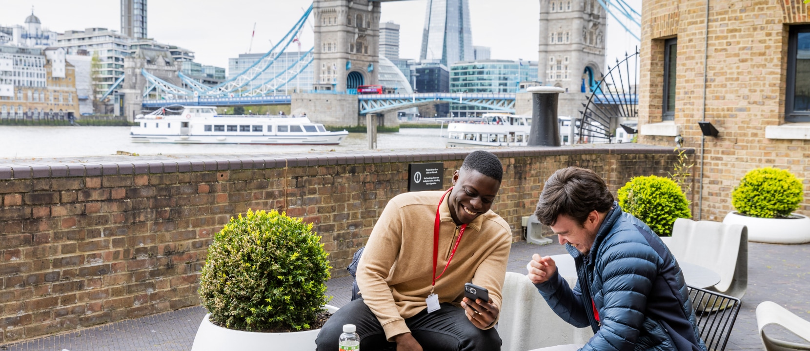 Two men are sitting outdoors and laughing while looking at a phone. They are near a brick wall, with Tower Bridge and a boat on the river in the background. Green plants are placed around their seating area.