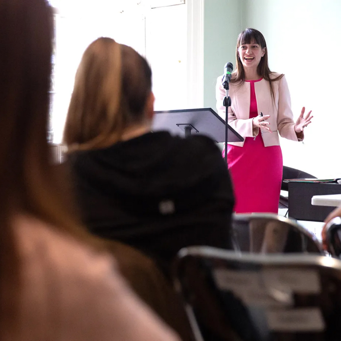 A woman in a pink dress and jacket stands at a podium, speaking into a microphone. She is engaging with an audience seated in front of her in a bright room.