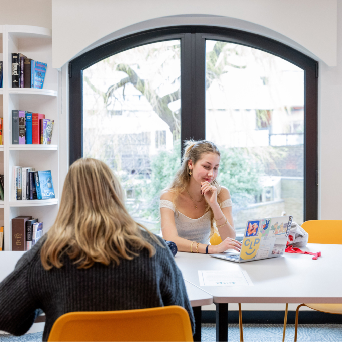 Two people are studying in a library with large windows. One person is using a laptop with stickers, and the other is facing away, seated at a table. Bookshelves are visible to the left. Bright and calm atmosphere.