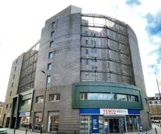 A modern, multi-story building features a Tesco Metro store on the ground level. The structure has a curved facade with large windows and brickwork. The sky is partly cloudy, adding to the urban setting.