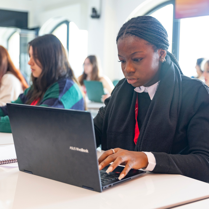 A focused person is typing on a laptop in a bright classroom, surrounded by others working at desks. The atmosphere is studious and collaborative.