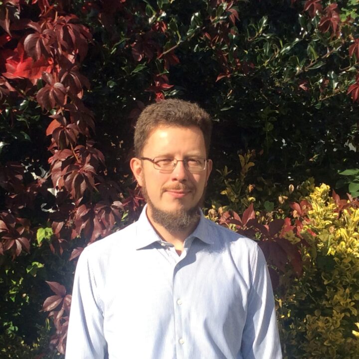 Dr. Lars Kjaer, a man with a beard and glasses, stands outdoors against a backdrop of vibrant red and green foliage. He's dressed in a light blue button-up shirt and smiles slightly as sunlight casts gentle shadows on the plants around him.