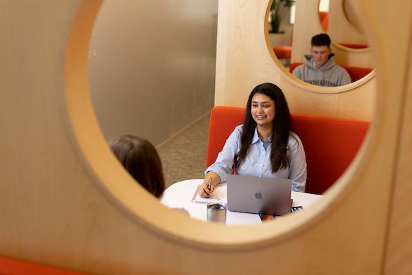 A woman with long dark hair sits at a table in an orange booth with a laptop, smiling at a person opposite her. In the background, a second individual in a gray hoodie is seated. The scene is framed by a circular wooden opening.