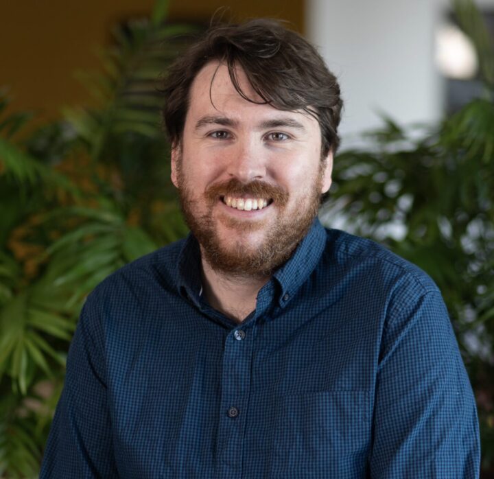 Dr. Tim Davis, sporting a beard and brown hair, smiles warmly at the camera. He’s clad in a blue checkered shirt, standing amidst lush green plants indoors, his approachable demeanor perfectly complementing the vibrant surroundings.