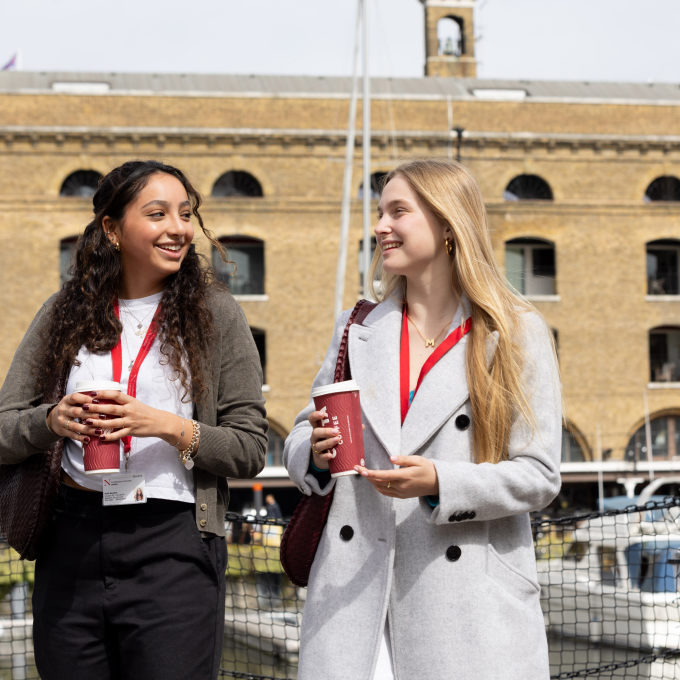 Two people stand outside a building, smiling and holding coffee cups. They are wearing ID lanyards and coats. The background features an industrial-style structure with large arched windows and a visible ship mast.