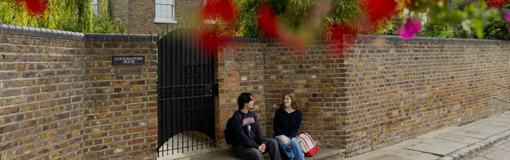 Two people sit on a sidewalk near a brick wall with a "Dockmaster's House" sign. They face each other, surrounded by greenery and red flowers. The scene is calm and casual, capturing a moment of conversation.