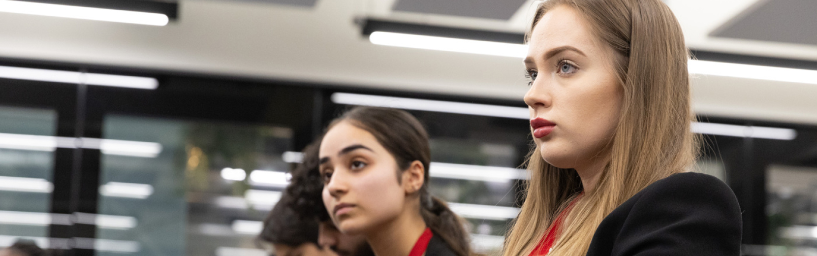 Two women attentively listening in a modern, brightly lit room. The woman in the foreground has long hair and wears a black blazer, while the woman in the background has dark hair tied back and wears a red shirt.