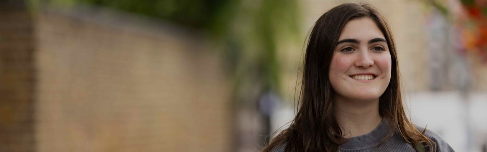 A young woman with long dark hair is smiling while standing outdoors. She is wearing a gray shirt, and behind her is a blurred background of a brick wall and green foliage.