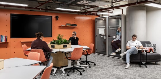 A modern office space featuring orange walls and gray carpet. Four people work independently: one at a round table with a plant centerpiece, one in a phone booth, another sitting on a sofa, and the fourth at a separate desk with a TV screen nearby.